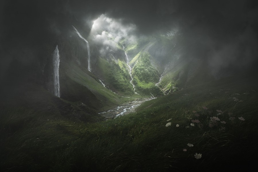 A view up a narrow, steep valley, with multiple waterfalls, overcast by dark clouds.