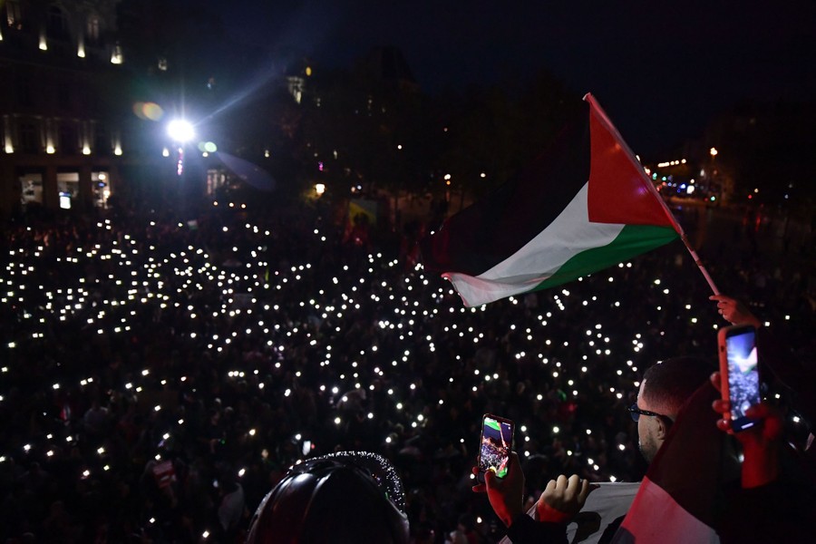 Protesters gather in a city square at night, shining the lights on their phones.