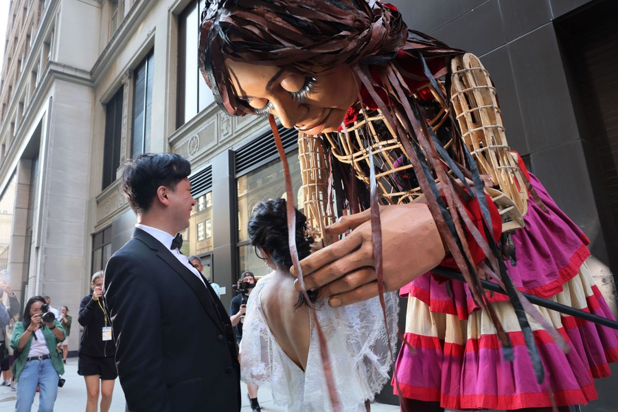 A large puppet of a girl leans over to embrace a newlywed couple.