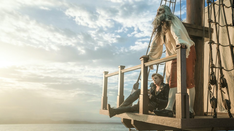 Rhys Darby and Taika Waititi on a ship with a bright cloudy sky behind them