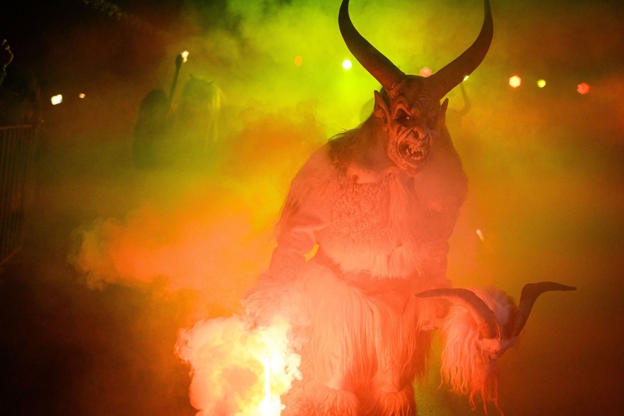 A person wearing a scary shaggy-horned-demon costume walks through smoke in a parade.