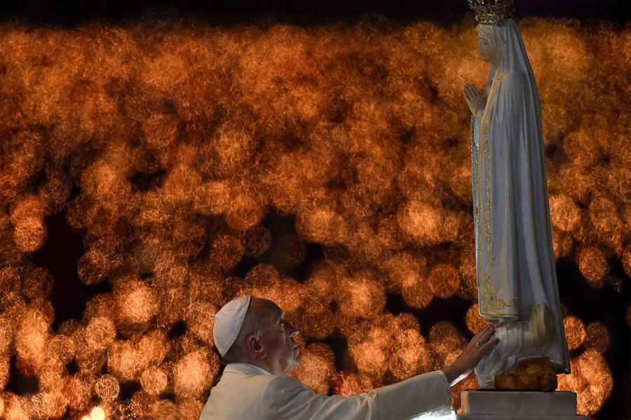 Blurred handheld candles burn in the background as Pope Francis touches a statue.