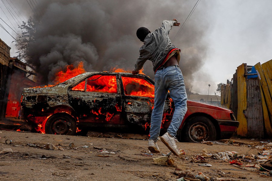 A protester throws stones over a burning car, toward riot police.
