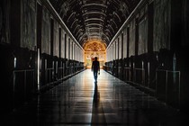 Photo of a figure holding keys walking down a long, dark aisle toward a frescoed, lit door in the distance