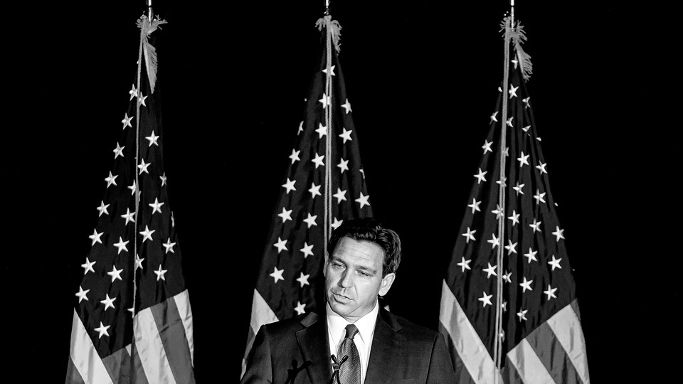 Black-and-white photo of Ron DeSantis speaking into a microphone, in front of three American flags