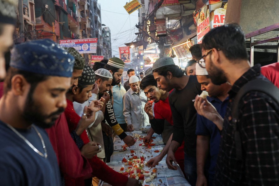A group of men eat food together, standing beside a long table in a street.