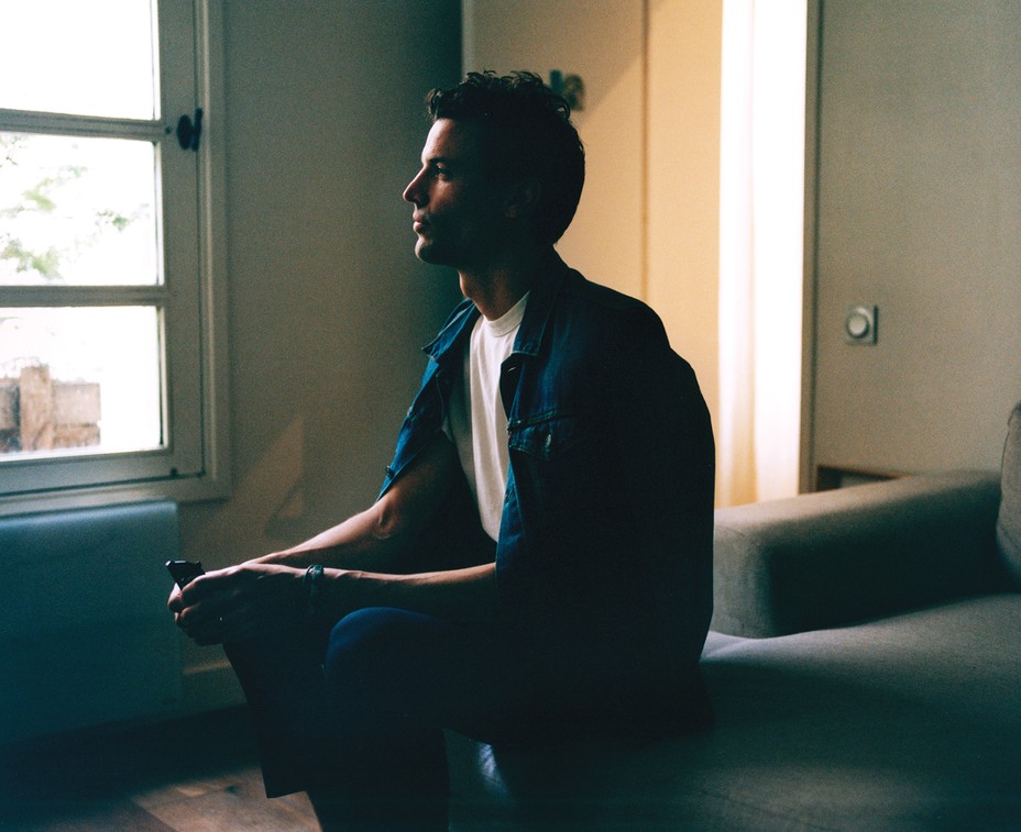 photo of man sitting sideways in blue jacket with window and door behind