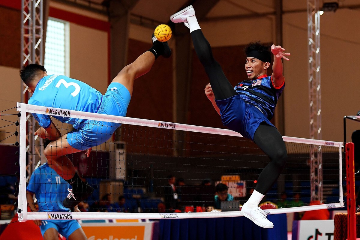 Two athletes leap high into the air to kick a small ball over a net.