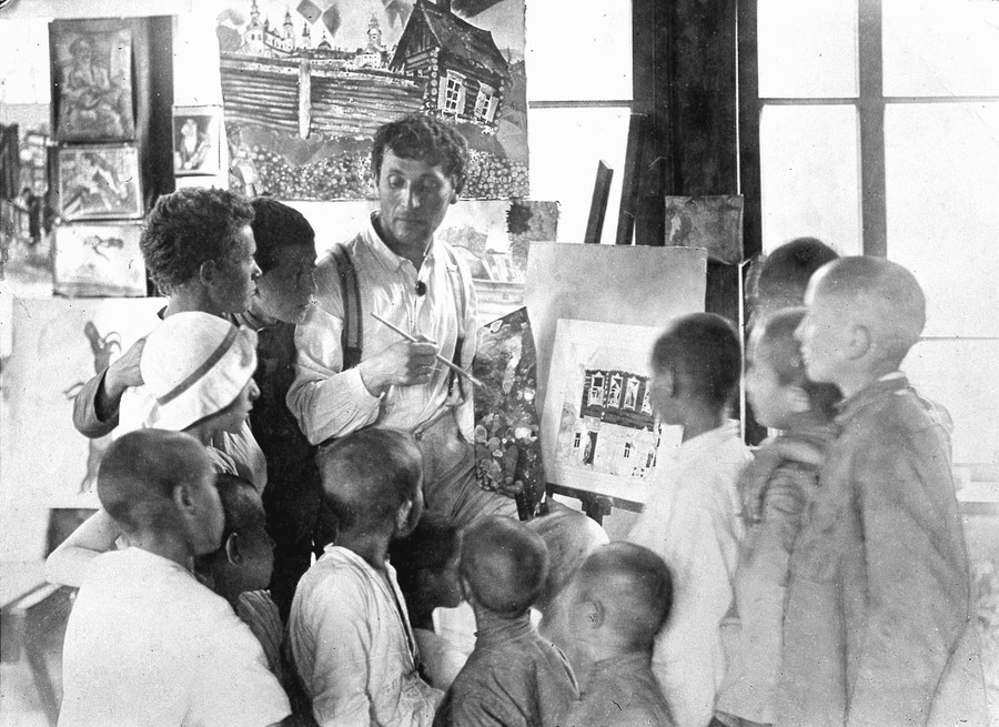 A man works on a painting, surrounded by a dozen children.