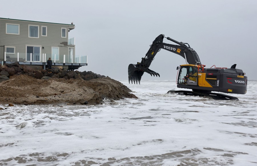 An excavator operates beside a beach house, digging while parked in the surf, in about three feet of water.