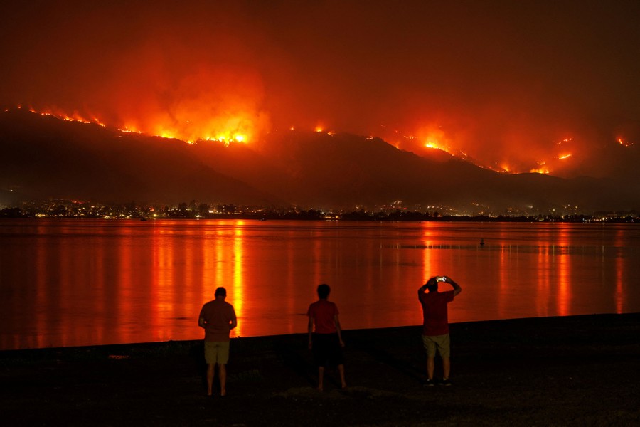 People stand on a lakeshore at night, looking toward wildfires burning across mountain ridges.