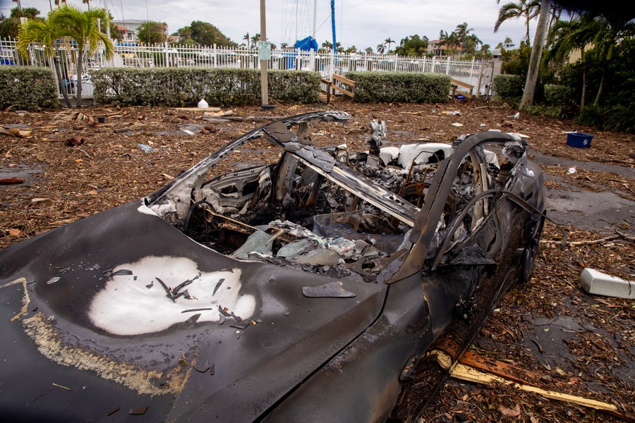 The burned-out shell of a car sits in a debris-strewn parking lot.