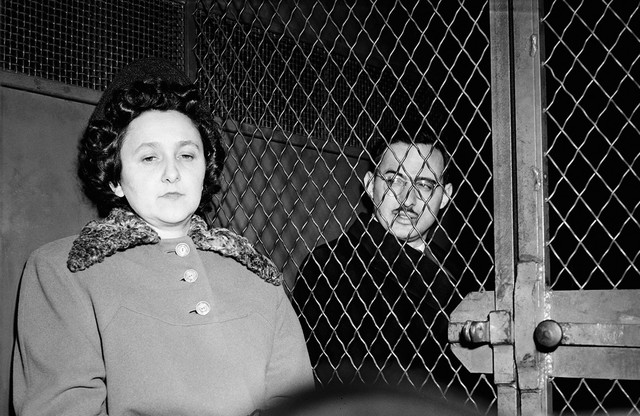 black-and-white archival photo of woman with eyes downcast, in hat and coat with fur collar, sitting next to man looking at her, with dark hair, glasses, and mustache, with a lattice-wire locked barrier between them