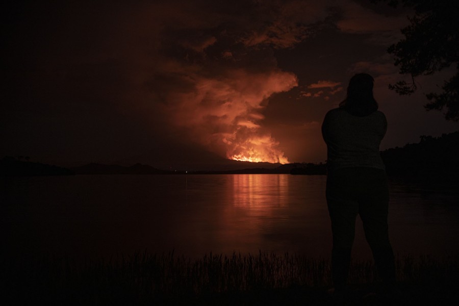 A person stands near the shore of a lake at night, observing a glow and rising smoke from a lava flow in the distance.
