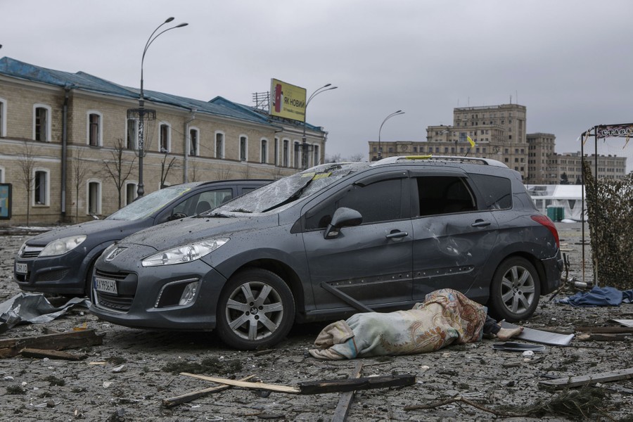 The blanket-wrapped body of a person lies on the ground beside damaged vehicles and among scattered debris.