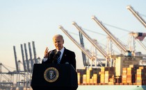 Joe Biden speaking from a lectern in a port, stacks of shipping containers visible on the background.