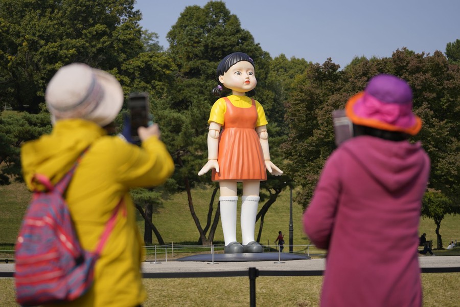 People photograph a tall statue of a young girl doll in a park.