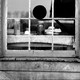 A black and white photo of a window in a wooden house, showing a table inside with jars on it