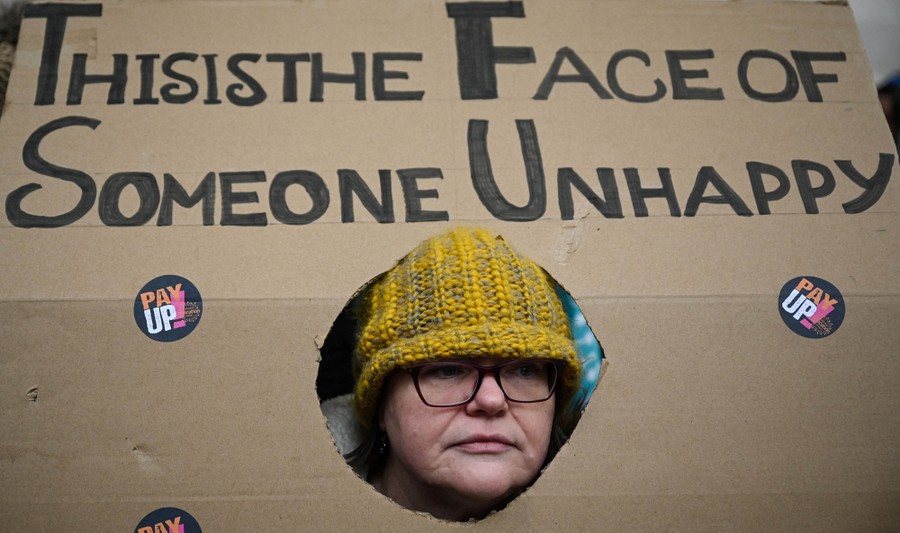 A protester pokes their face through a hole in a cardboard sign that reads "This is the face of someone unhappy."