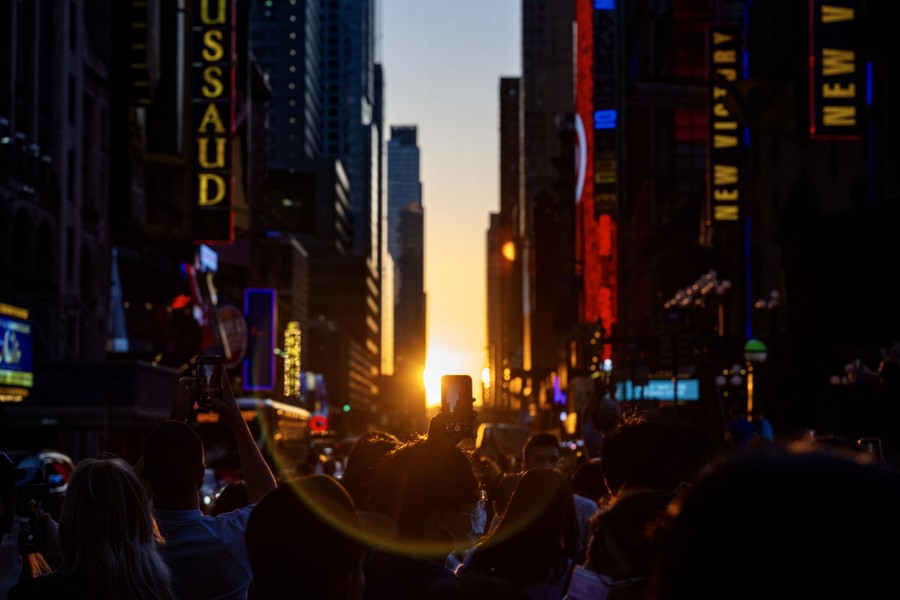 Dozens of people stand in a Manhattan street, holding up their phones and looking toward the setting sun between buildings.