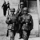 Black-and-white photo of four Nazi soldiers walking through a city street with rifles strapped to their backs as a policeman watches