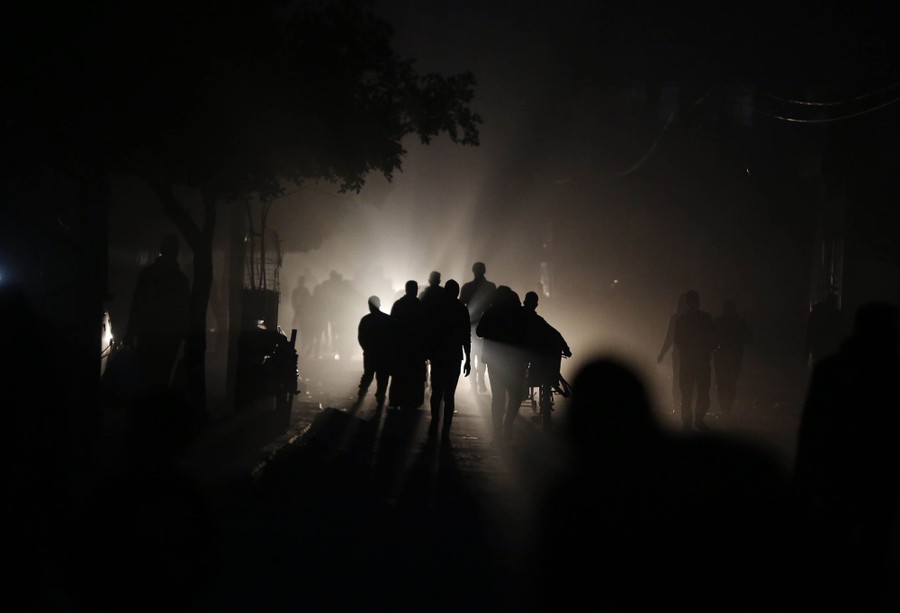 Crowds of people carry belongings down a dark street, backdropped by a distant light.