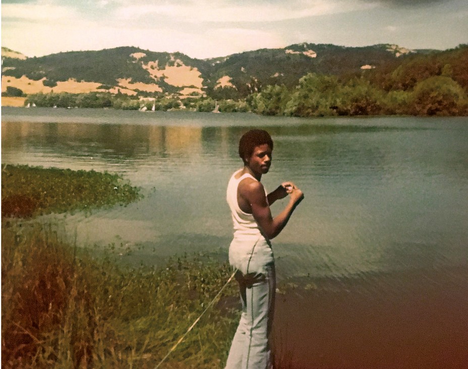 a man at a lake looks at the camera holding a fishing surrounded by mountains circa 1970s.