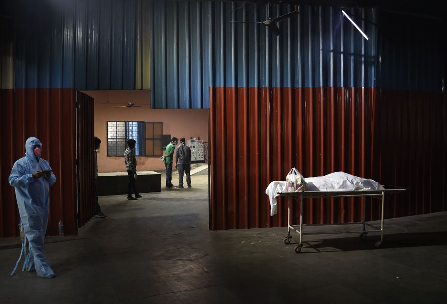 A worker wearing protective gear stands near a single body, covered in a sheet, on a carrier, inside a crematorium.