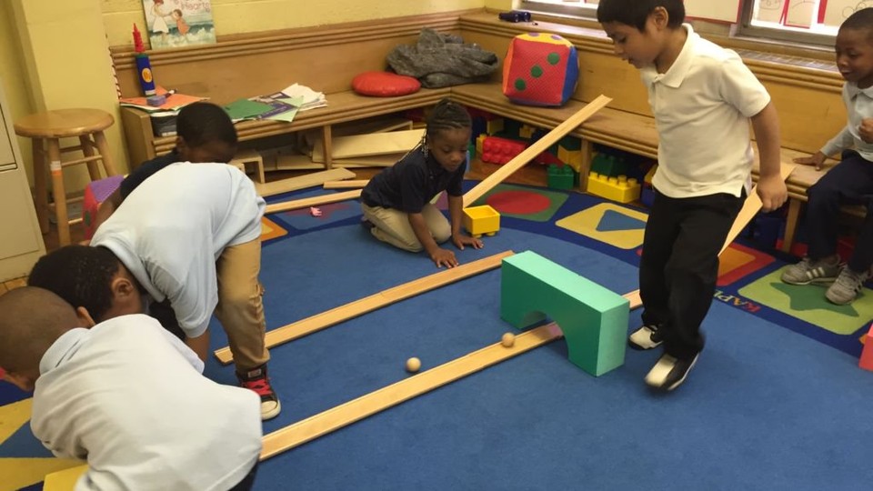 A group of pre-school students play on a blue carpet with wooden ramps and balls.