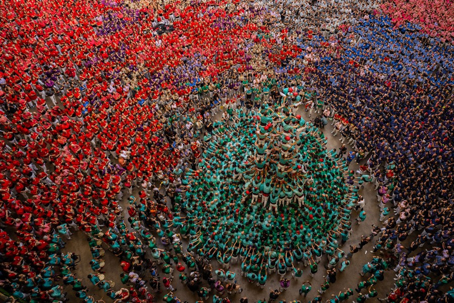 A large crowd of competitors and onlookers fill an arena space, looking up at a troupe that is forming a tall human tower.