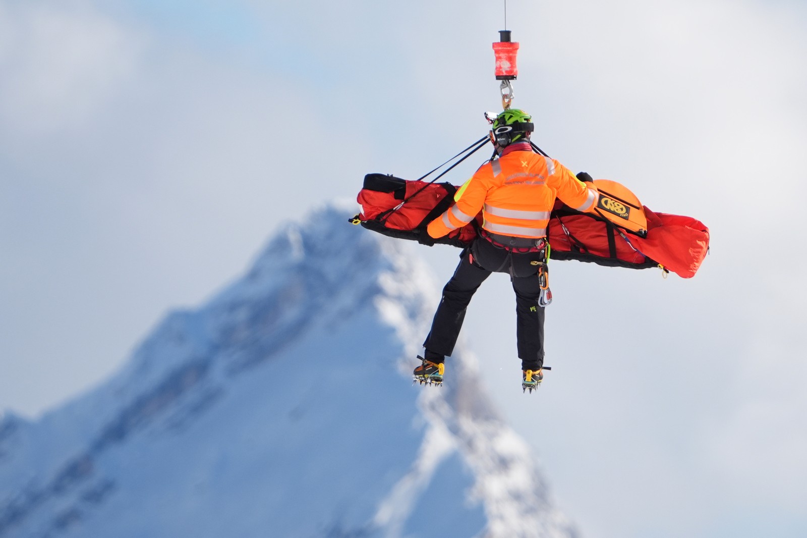 A rescue worker hangs beside a covered stretcher that carries an injured athlete, both suspended beneath an unseen helicopter.