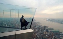 A person dressed as Batman stands on a high skydeck above the New York skyline.