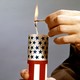 A person's hands holding a red-white-and-blue firecracker while lighting the wick with a match