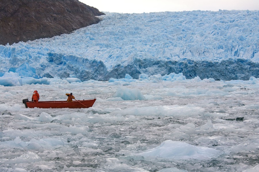 Two people pilot a small boat through floating ice at the terminus of a glacier.