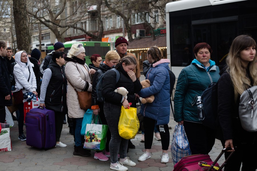 A line of more than a dozen people stands on a city sidewalk, waiting for a bus.