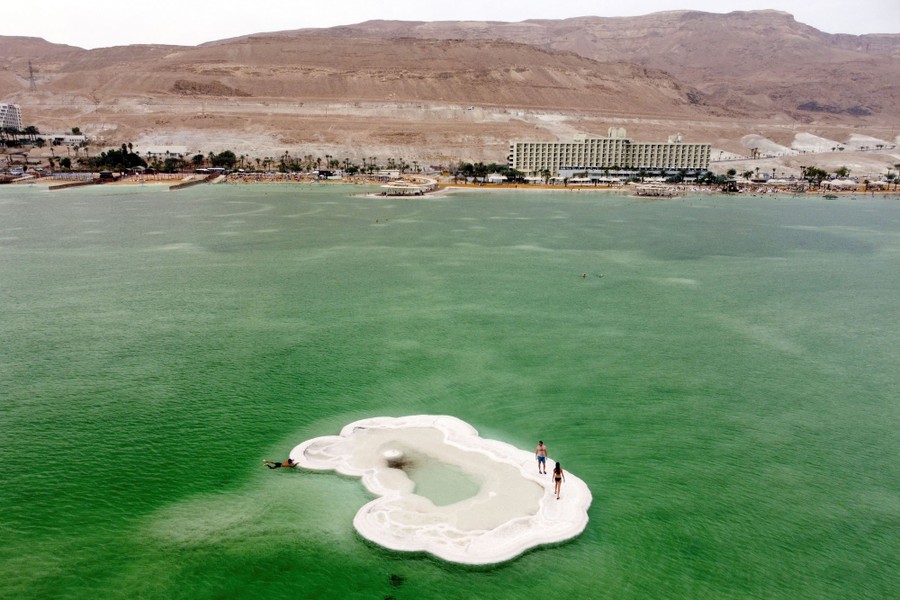 Two people walk on a small formation of white salt in the midst of the green water of the Dead Sea.