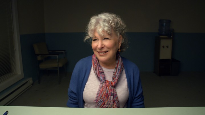 The actor Bette Midler sits at a desk wearing a white shirt and a blue sweater