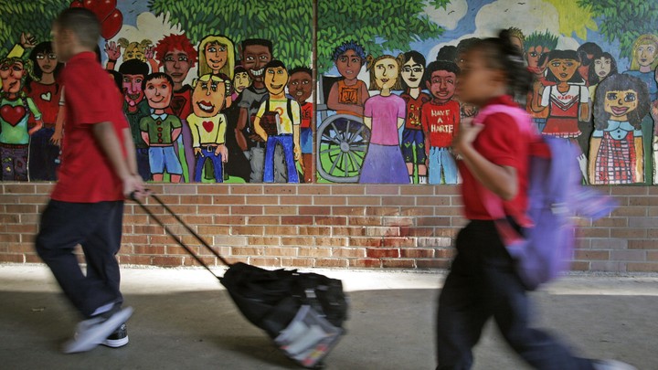 Two black children walk with backpacks past a mural depicting a diverse array of children.