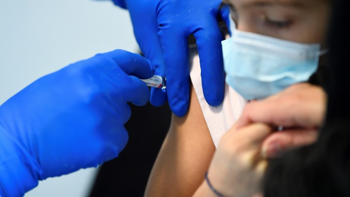 A pair of hands in blue gloves administer a vaccine to a young person's arm.