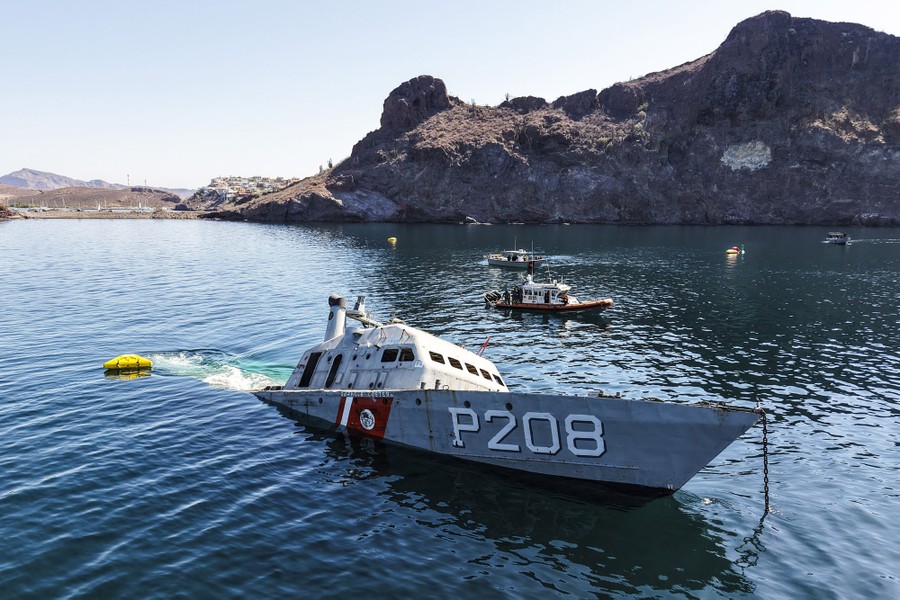 An aerial view of a small navy ship being sunk near a rocky coastline.
