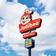 photo of roadside red Jollibee sign with smiling bee, Drive-Thru, and open 24 hours on it, with bright blue sky and clouds in background