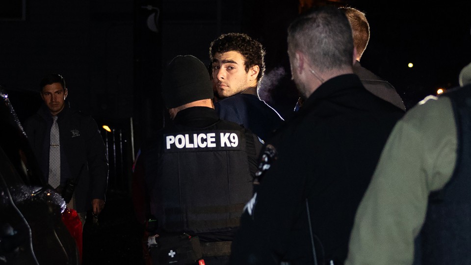 Photo of Luigi Mangione after his arrest looking back at the camera while being escorted away by a cop in a “Police K9” vest