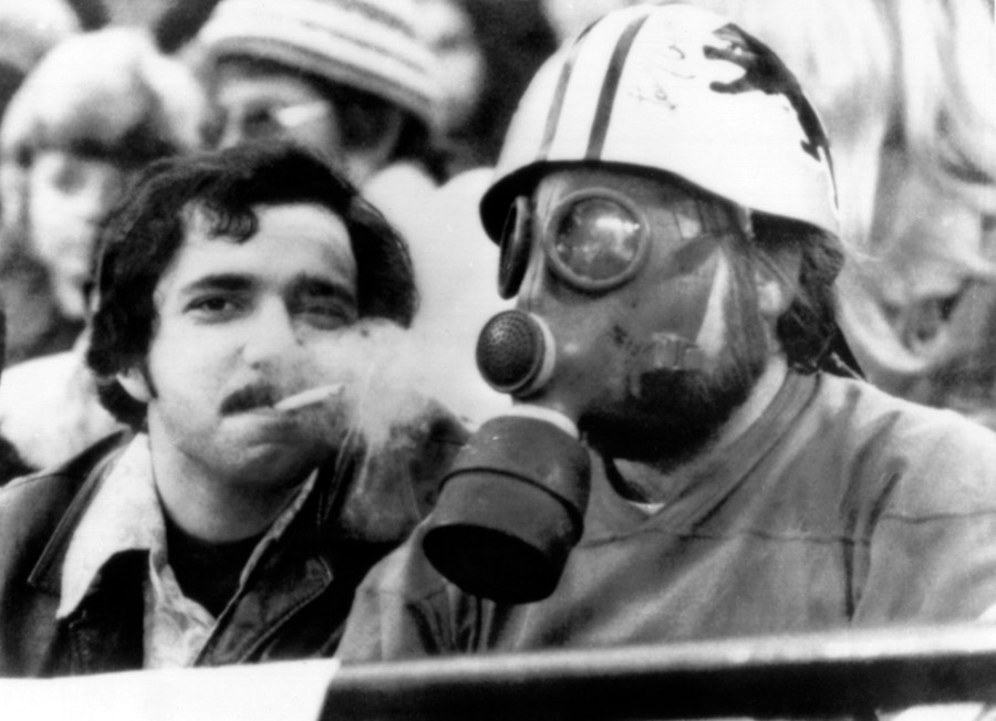 A football fan wears a helmet and a gas mask as another fan blows cigarette smoke toward them.