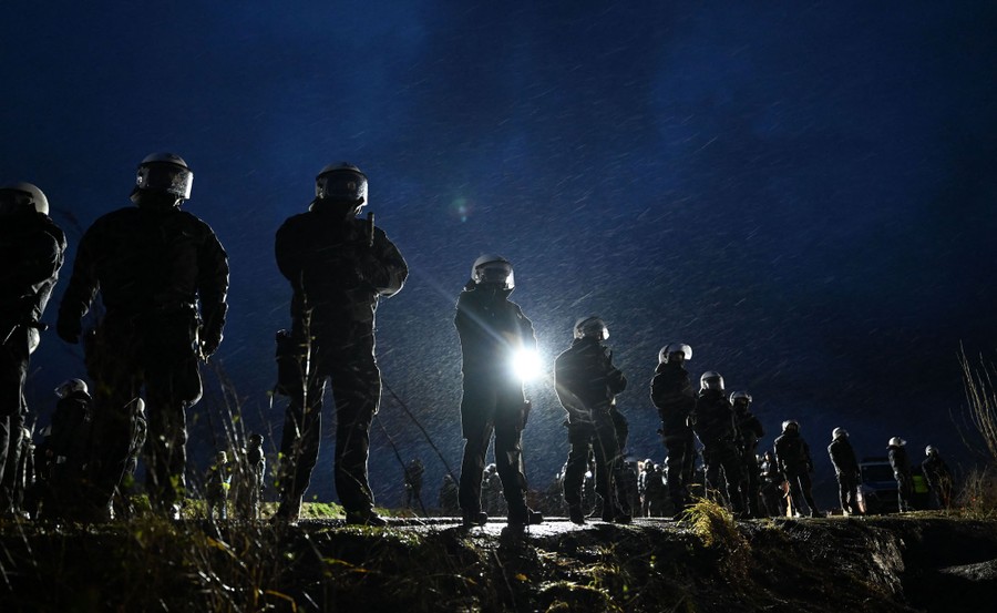 Riot police stand in a line on a road on a rainy night.