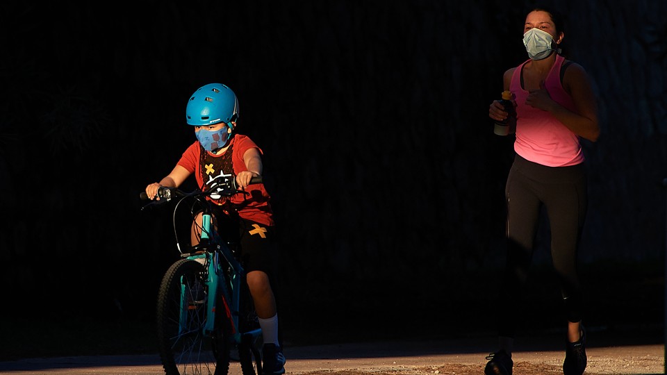 A child wearing a mask rides a bike in front of a masked jogger.