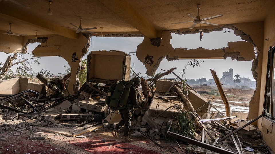 A soldier walks through a blown out interior in Gaza, ceiling fans and carpets visible under rubble but walls blasted through.