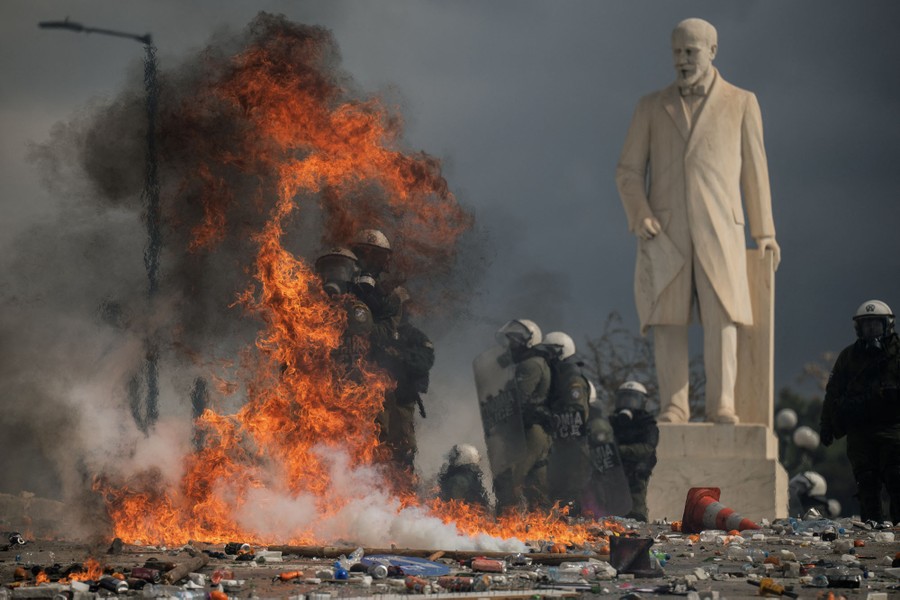 Riot police officers stand in full gear beside a statue, near flames and debris in a street.