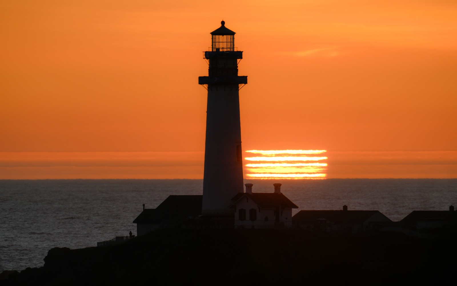 The sun sets behind a lighthouse, with clouds and refraction separating the sun into about six stripes on the horizon.