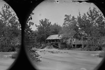 A black-and-white photograph of a house with a stream running near it, surrounded by trees and water