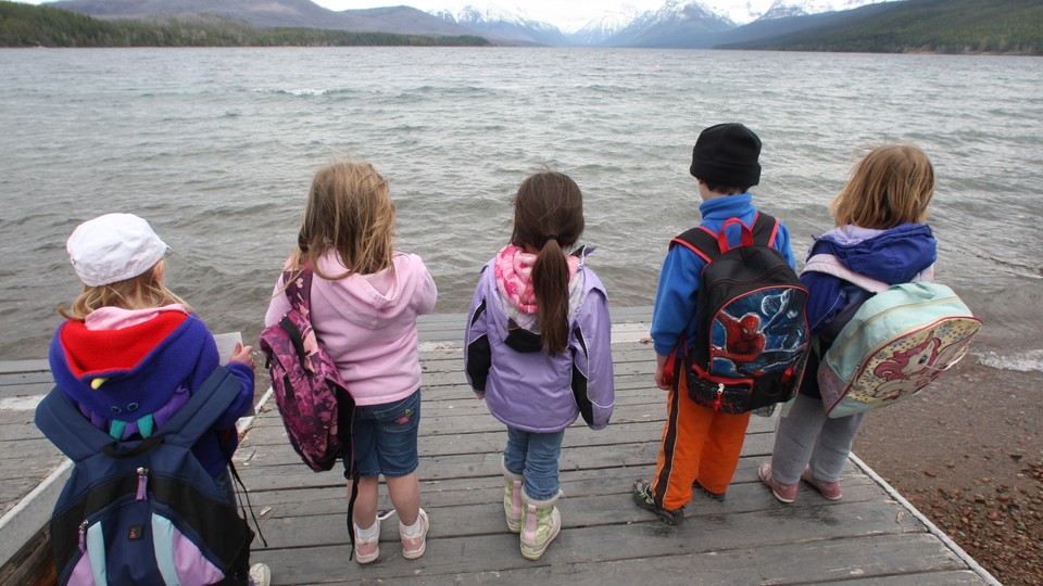 Five children holding backpacks look out at the expansive lake and mountains of Glacier National Park.